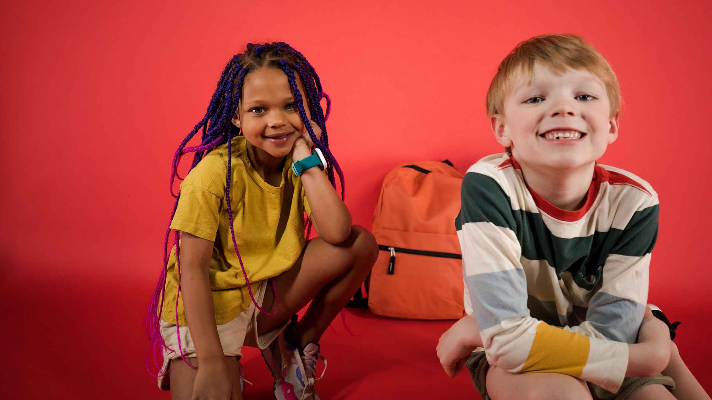 Two young children smile at the camera with a backpack sitting behind them